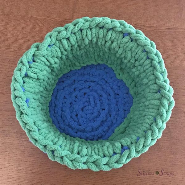 The inside of a round basket, with a blue bottom and green sides, on a wood table. Learn how to knit without needles! This bright, finger knit basket pattern is fast and easy to make. It stands up tall with the help of some plastic canvas hidden between the layers. Find the free pattern on Stitches n Scraps.