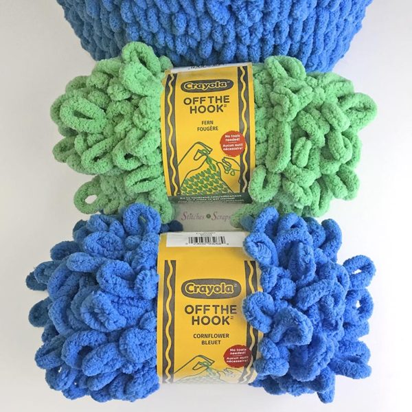 Two skeins of Crayola off the Hook yarn from Lion Brand. One in Fern Green and one in Cornflower Blue. Pictured on a white table, in front of a basket made from this yarn. Finger Knit Basket - Pattern on Stitches n Scraps