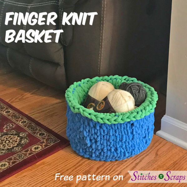 A fuzzy, finger knit basket in bright blue and green, on a wood floor next to a beige wall, brown couch, and red rug. Basket is filled with skeins of yarn. Finger Knit Basket - Pattern on Stitches n Scraps