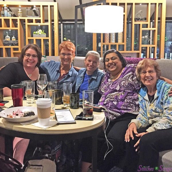 5 women on a couch in a hotel lobby, with a round table in front of them and wooden shelves behind them. The table is filled with empty glasses and plates. 
