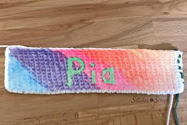 A crocheted swatch, with a diagonal rainbow across it, surrounding the name Pia written in green. 