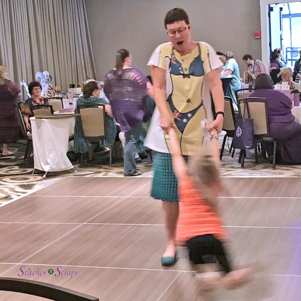 An adult woman, twirling a small child on a dance floor with people seated in the background.