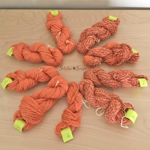 9 mini skeins of yarn, numbered with green tags and arranged in a circle on a wood table. Half the yarn is solid orange, the other half is orange plied with dark brown thread.