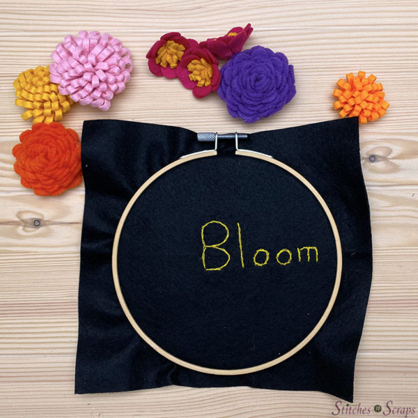 Square of black felt in an embroidery hoop, with the word Bloom embroidered on it. Several felt flowers nearby. 
