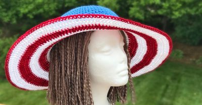 Summer Glory Sun Hat - a free pattern on Stitches n Scraps. Sun hat with red and white striped brim and blue crown on a foam head with brown yarn "hair". Head is placed on a wood railing overlooking trees and grass.
