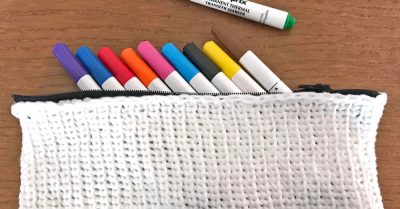 White, crocheted pencil case with a black zipper, on a wood table. Pencil case is full of markers in different colors. Back to School Pencil Case from Stitches n Scraps