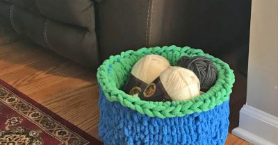 A fuzzy, finger knit basket in bright blue and green, on a wood floor next to a beige wall, brown couch, and red rug. Basket is filled with skeins of yarn. Finger Knit Basket - Pattern on Stitches n Scraps