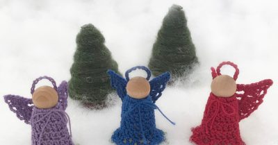 4 peg doll angels with lavender, blue, and red crocheted dresses and wings, in front of yarn wrapped fir trees on a snowy background. Wording says "Peg doll angel. Free crochet pattern on Stitches n Scraps