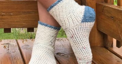 Two feet, standing crossed on a wood deck with a wood rail. The feet are wearing white socks with blue toes, heels and cuff edging. Wording reads Walking on Clouds - a free crochet pattern from Stitches n Scraps