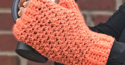 hands wearing orange and black, crocheted fingerless mitts, and holding a travel coffee mug. A red brick wall in the background and the words Autumn's Kiss a free crochet pattern on Stitches n Scraps
