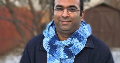 A man in a black jacket and blue scarf outdoors with light snow on the ground. Text says Still Waters Scarf - A free crochet pattern on Stitches n Scraps