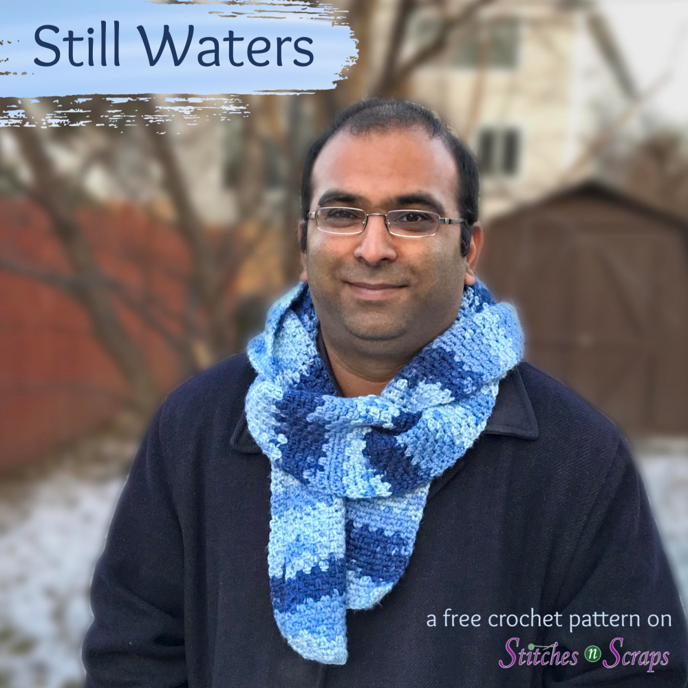 A man in a black jacket and blue scarf outdoors with light snow on the ground. Text says Still Waters Scarf - A free crochet pattern on Stitches n Scraps